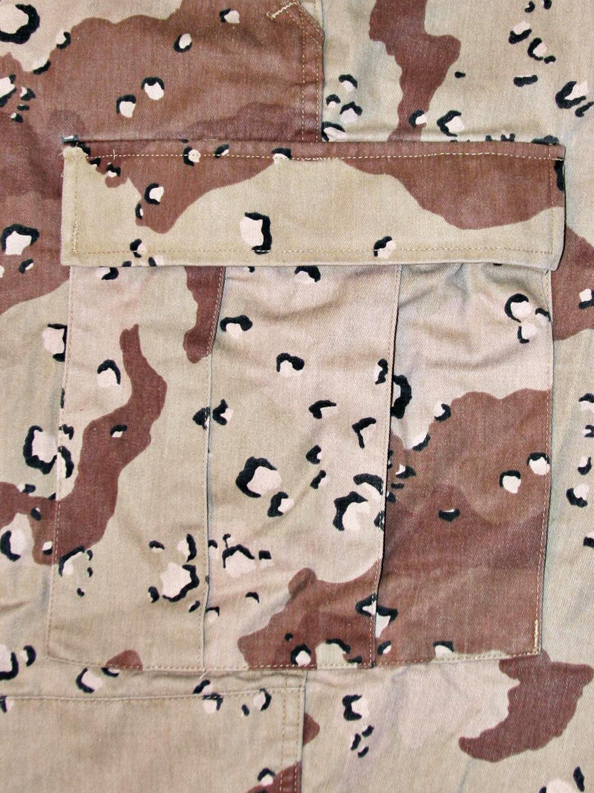 Close, flat view of the six-colour desert camouflage fabric of a real uniform garment: a pale sand/cream ground with rust-brown and dark brown shapes, and small black spots each sitting inside a pale off-white chip; a stitched seam and pocket edge are visible. Colour rendering depends on the light and the production run.