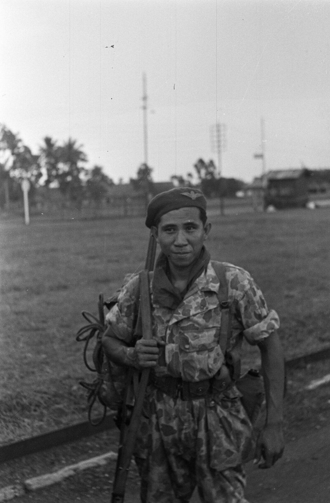 A paratrooper in camouflage uniform, wearing a beret and carrying a parachute pack and weapon, standing on an airfield in Indonesia in December 1948, with radio masts and palm trees in the background.