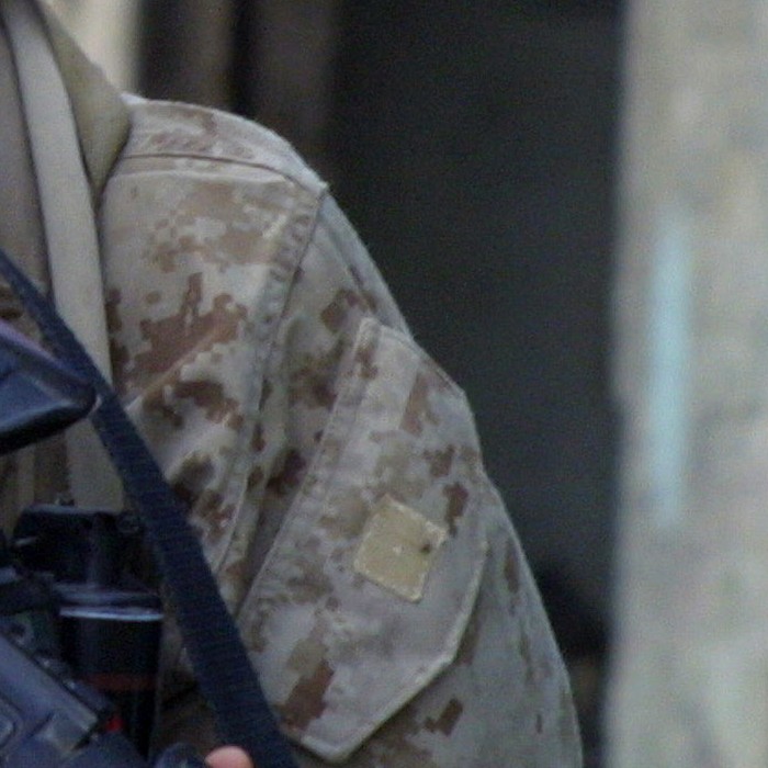 Close-up of sandy digital camouflage fabric with visible stitching and a pocket flap, showing rectangular pixel blocks in tan, sand and grey-brown.