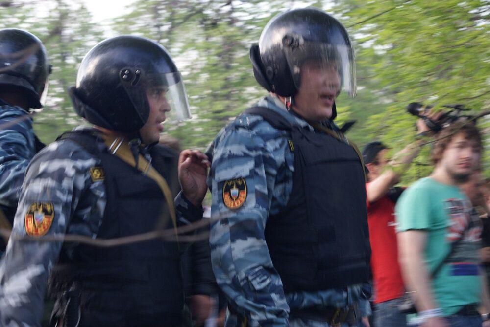 Police officers in blue-grey camouflage under black riot helmets and body armour, moving past a blurred crowd of onlookers and photographers.