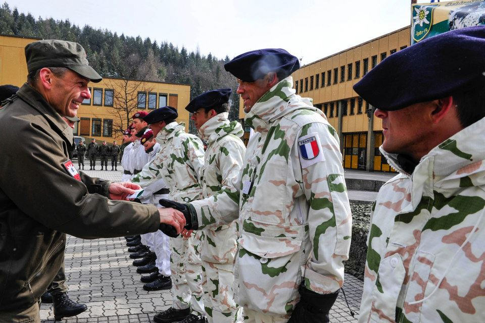 A line of soldiers in pale Tundra camouflage — white ground with green and salmon shapes — being greeted in a paved courtyard outside a barracks building, two of them wearing the wide dark Alpine beret and one with a French tricolour patch on the sleeve.
