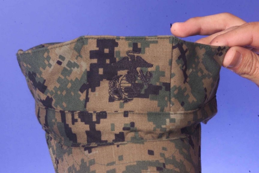 Close-up of a green, brown and black pixel-patterned cap cover held in a hand, with an embroidered Eagle, Globe and Anchor emblem on its front.