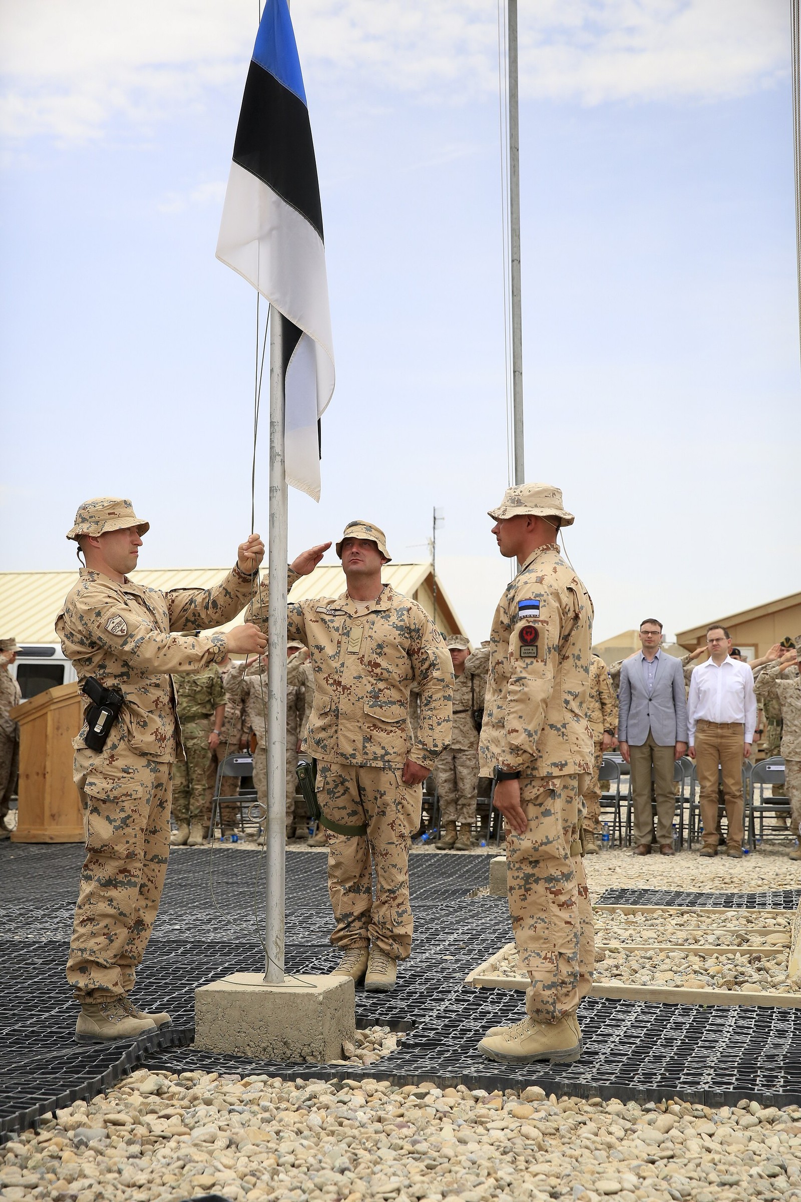 Three Estonian soldiers in desert pixelated camouflage with Estonian flag patches lowering the Estonian flag at a military ceremony.