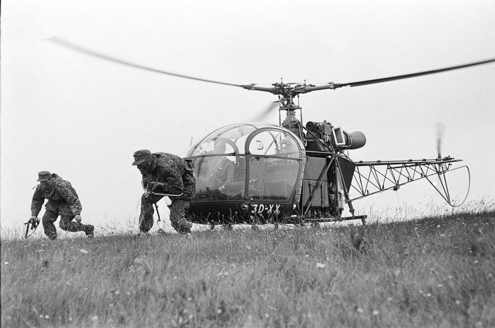 Two Austrian Bundesheer soldiers in Erbsentarn camouflage moving past an Alouette II helicopter during a military academy exercise at Ramsau-Hainfeld, 1963.
