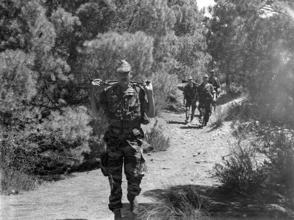 French paratroopers in camouflage uniforms walking in single file along a track through scrub in Algeria, 1961