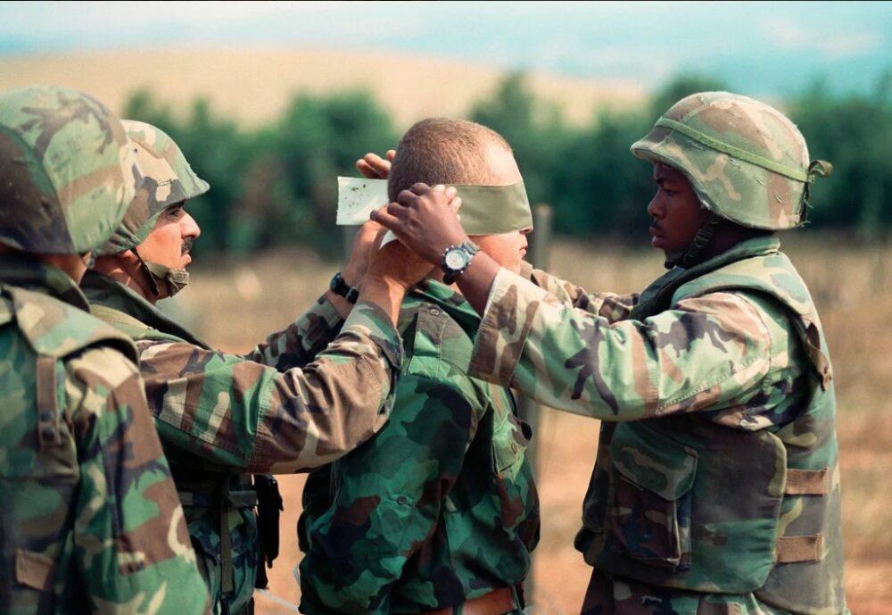 Yugoslav soldier detained in Kosovo in July 1999, blindfolded by a U.S. Marine before transport, wearing a green-dominant large-leaf oak pattern camouflage jacket of the Yugoslav M89 family