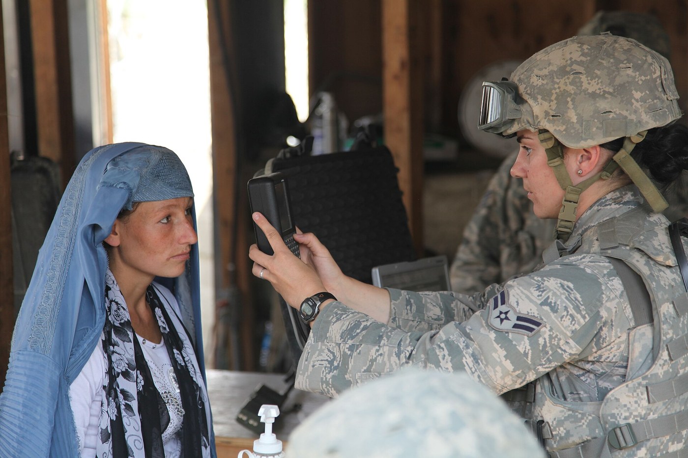 An airman in an Airman Battle Uniform and body armour scans the iris of an Afghan woman with a handheld device in the waiting area of a hospital.