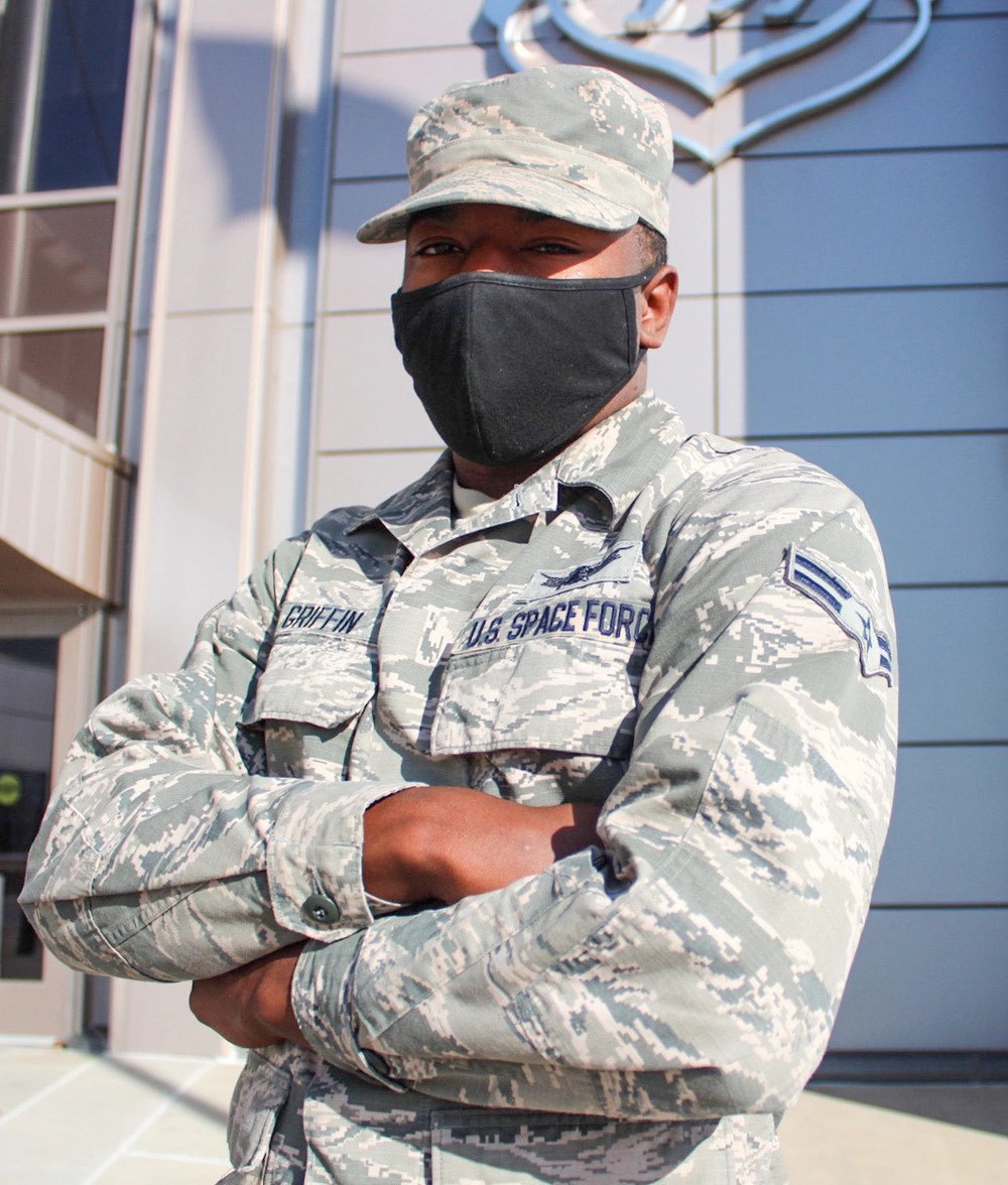 A Space Force airman in an Airman Battle Uniform with a U.S. Space Force name tape and a black face covering, standing outside a building.