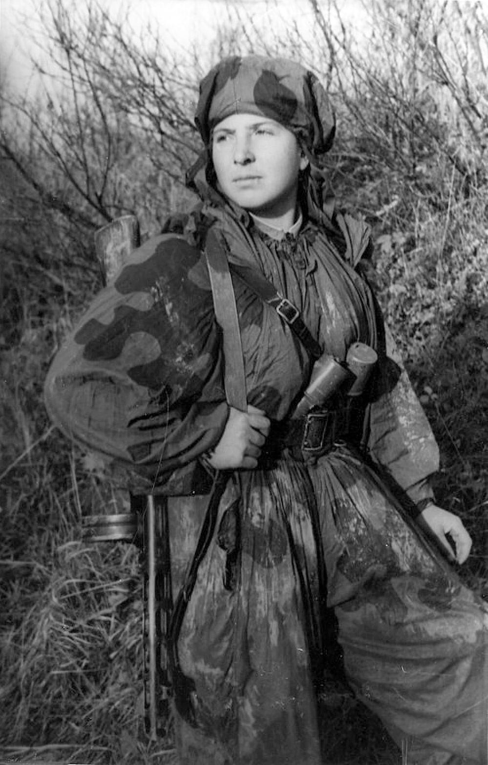 Soviet woman soldier in a hooded two-tone camouflage smock printed with large dark blobs, a submachine gun at her side, standing in brush