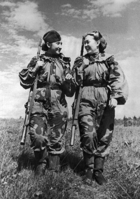 Two Soviet snipers in one-piece camouflage coveralls printed with large dark blotches on a light ground, rifles slung over the shoulder, walking along a path between bushes