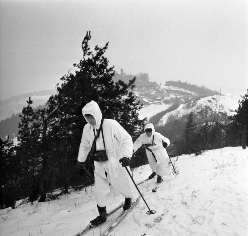 Two men in white two piece winter camouflage suits with hoods skiing downhill on a snow covered slope with trees and a castle in the background