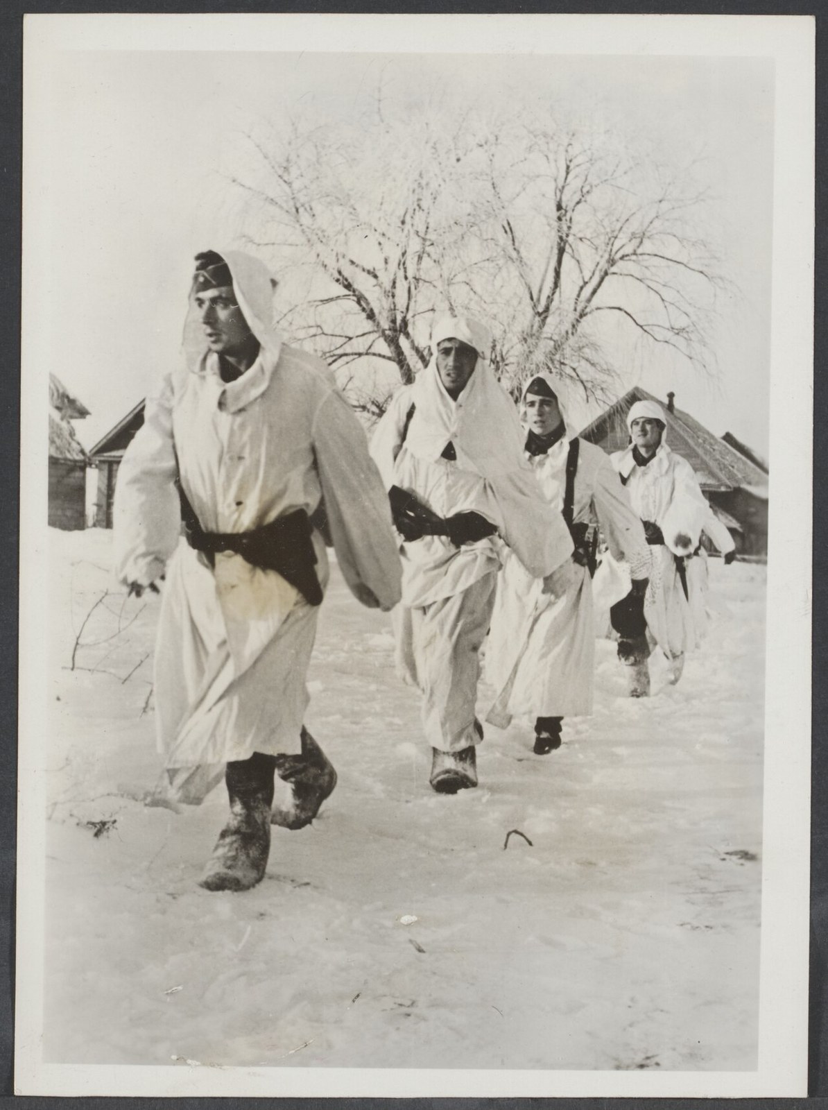 Four soldiers in long white snow camouflage smocks walking through a snow covered village street past wooden houses