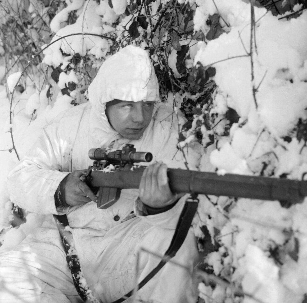 Sniper of the 6th Airborne Division wearing a hooded white snow camouflage suit with a scoped rifle, in snow covered ground